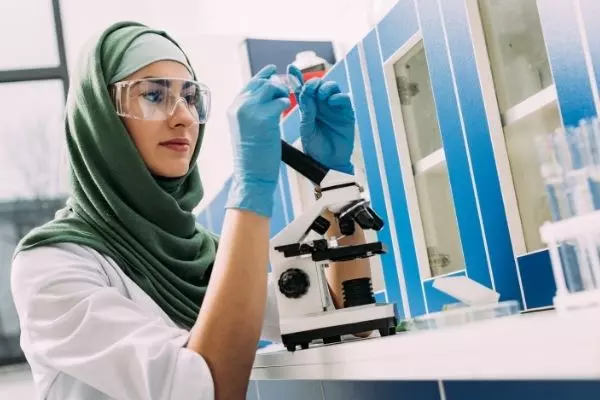 Una mujer con un pañuelo en la cabeza y gafas protectoras trabaja en el microscopio